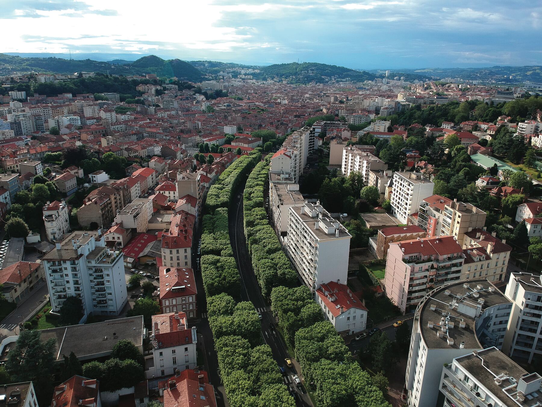Vue aérienne de Saint-Étienne : une avenue bordée d'arbres traverse un quartier d'immeubles aux toits rouges, avec les collines au fond