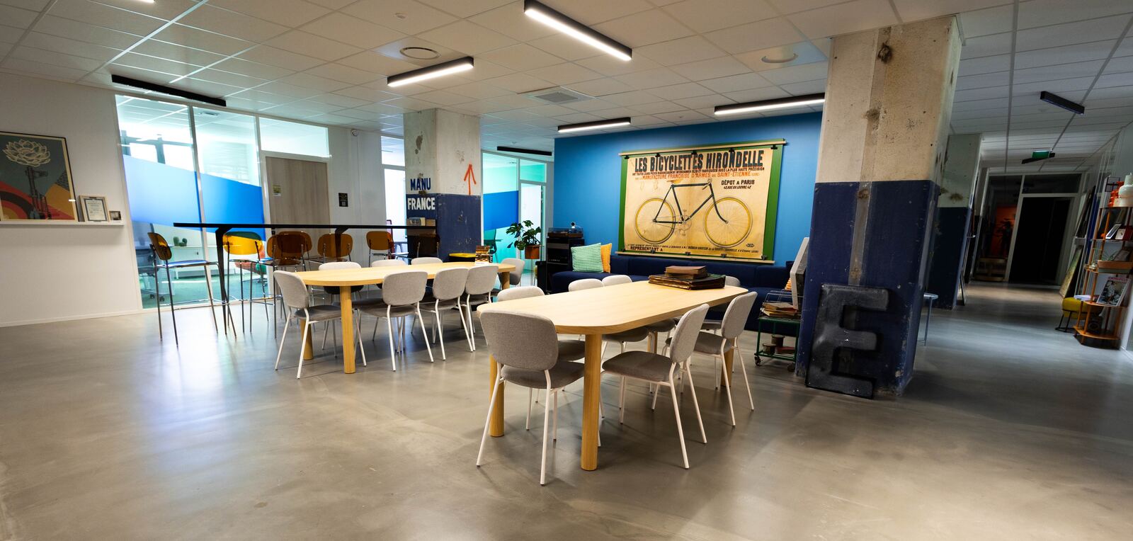 Salle commune d'un site de bureaux opérés : grandes tables en bois, chaises grises et affiche ancienne de bicyclettes